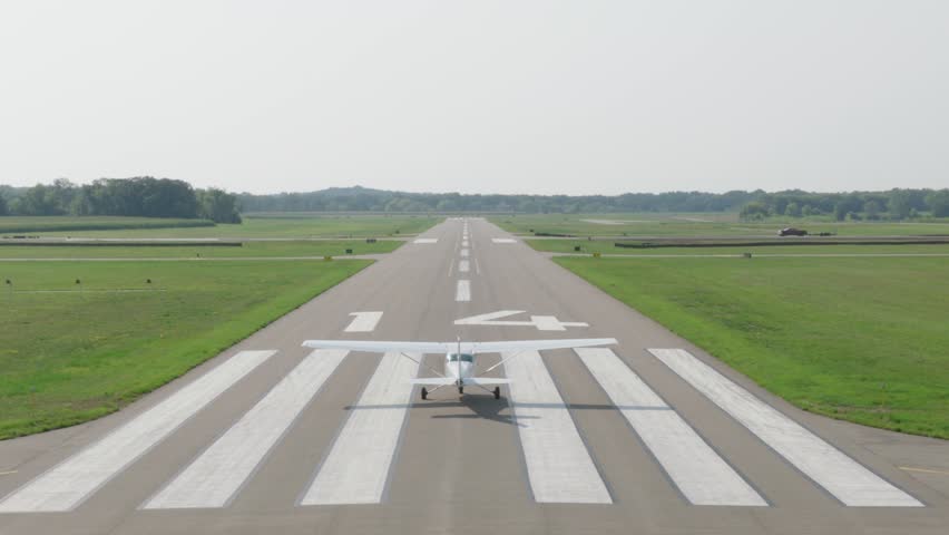 Small single engine airplane accelerates down empty runway and climbs into hazy sky above green countryside on calm summer morning
