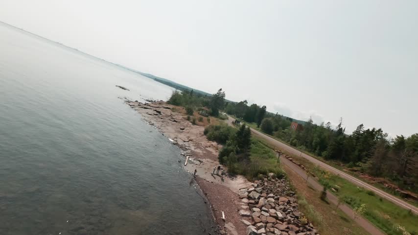 FPV drone skims rocky Lake Superior shoreline past trees and bridge near Lester River mouth in Duluth Minnesota on hazy summer day