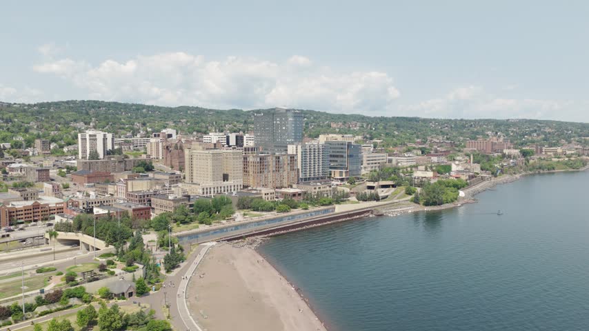 Drone orbits Duluth Minnesota skyline showing downtown buildings hillside neighborhoods and sandy beach along Lake Superior waterfront on clear summer day
