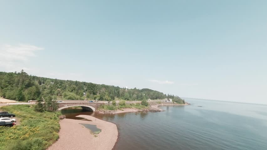 FPV drone glides past arched stone bridge over Lester River and rocky Lake Superior shoreline near Duluth Minnesota on hazy summer day