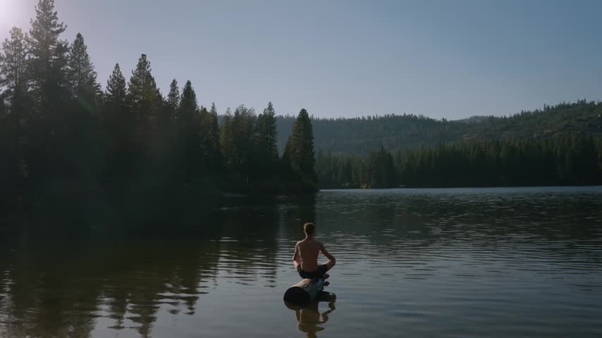 A man wades into a calm lake with a stand up paddleboard beside a pine forest shoreline.
