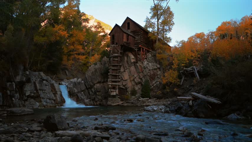 Scenic landscape footage showing historic wooden mill structure above cascading waterfall. Vibrant orange fall foliage surrounds rocky riverbed with moving water in cinematic setting.