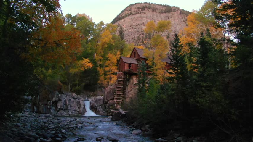 Scenic landscape featuring historical wooden mill perched on cliffs above rushing river and waterfall. Colorful autumn foliage surrounds crystal clear water under soft mountain light.