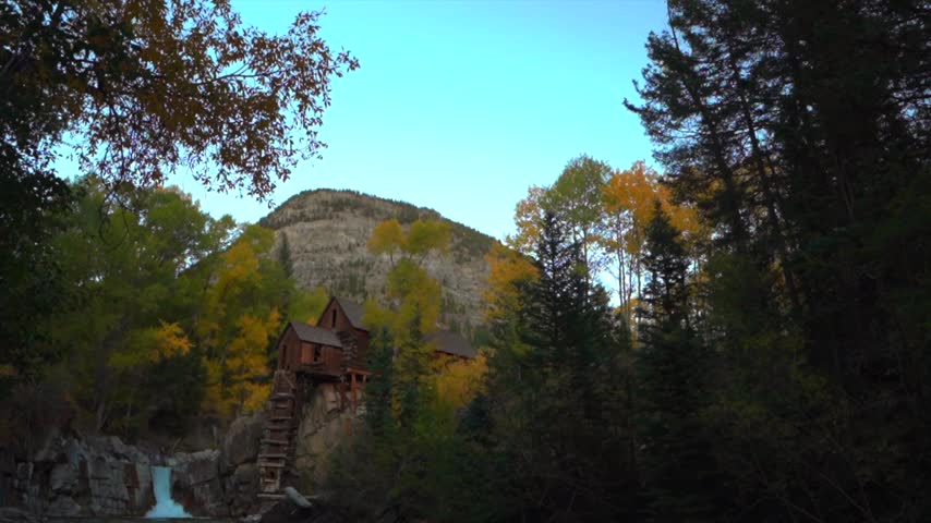 Slow camera pan revealing historic crystal mill nestled in a colorful autumn forest with a mountain backdrop, a serene waterfall, and clear river flowing through a rocky bed, capturing natural beauty.