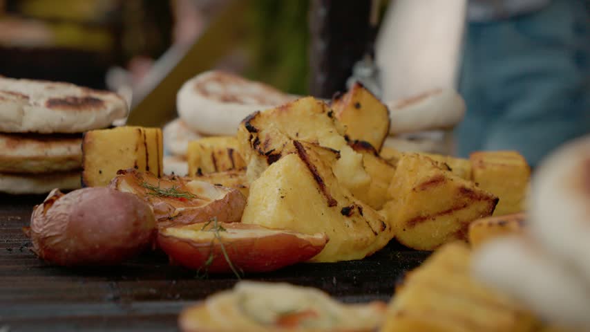 A whole baked potato is shown on a grill plate along with grilled vegetables at a street food festival. The event features various grill items for visitors.