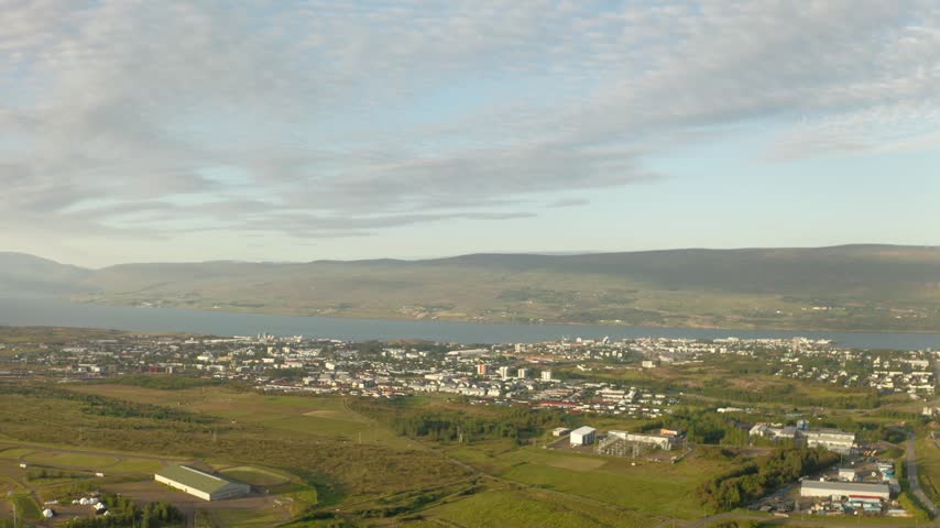 An aerial drone shot shows the city of Akureyri situated on the Eyjafjörður fjord in northern Iceland, surrounded by green landscapes and mountains.