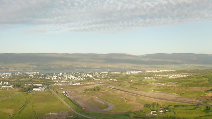 An aerial drone shot flies over a dirt racetrack on the outskirts of Akureyri, Iceland, with the city, Eyjafjörður fjord, and mountains in the background.