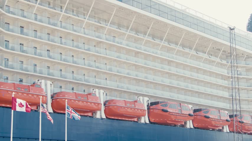 A static shot of a huge cruise ship docked in Akureyri, Iceland. The hull features rows of orange lifeboats and decks with glass balconies. International flags fly in the foreground.
