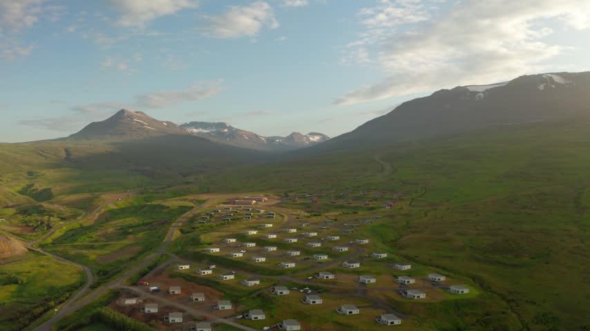 Aerial view of a village of small white cabins in a lush green valley, surrounded by majestic snow-capped mountains near Akureyri, Iceland during a sunny day.