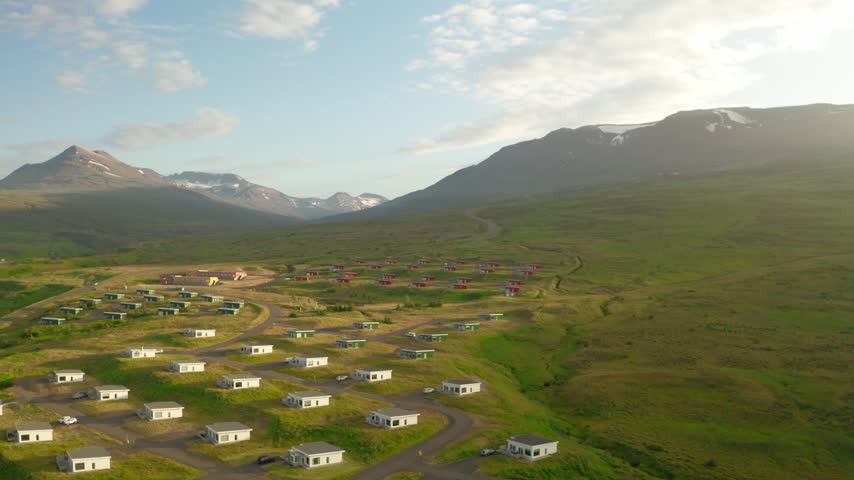 Aerial drone shot of a modern holiday village with colorful cabins in a green valley near Akureyri, Iceland. Majestic mountains with snow-capped peaks rise in the background at sunset.