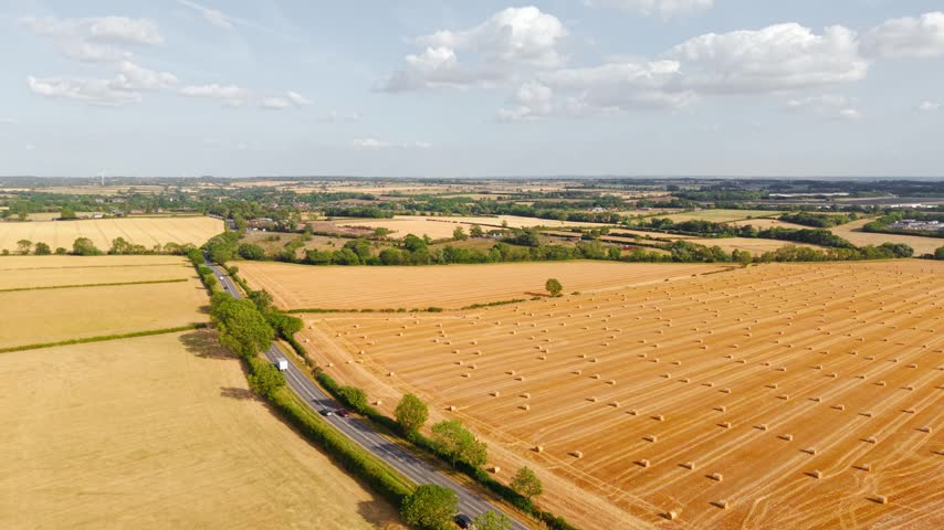 Hay bales aerial farmland with harvested wheat field and rural countryside. Round straw bales across agricultural landscape with country road under summer sky and scattered clouds. Livestock feed