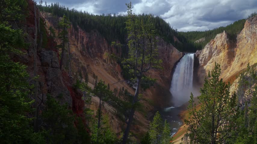 Scenic landscape video featuring lower falls of grand canyon of yellowstone national park with cascading water down rocky gorge surrounded by lush pine forest under moody cloudy sky.