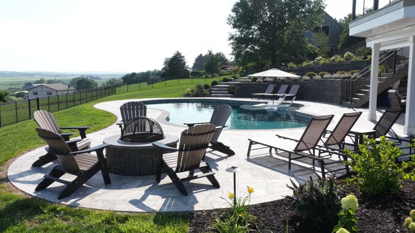 Curved swimming pool with stone patio overlooks green farmland valley in United States, wooden lounge chairs and fire pit sit beside covered deck under hazy summer sky. Aerial drone orbit shot