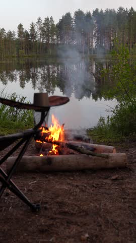 Burning campfire on the shore of a calm forest lake with a folding camping chair in the foreground at twilight. Concept of peaceful wilderness camping, evening outdoor relaxation, serene nature getaway and bushcraft lifestyle.