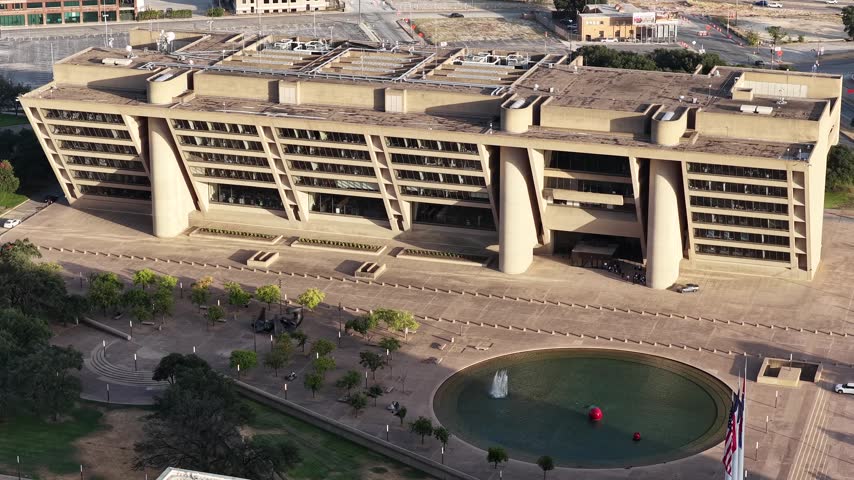 Aerial drone view of Dallas City Hall in the Government District, Dallas, Texas, at golden hour. Angled concrete floors step outward above deep round piers, throwing hard shade across the paving in the foreground. A circular pool with red spheres, flagpoles and rows of trees fill the open plaza below. A close orbit runs along the facade.