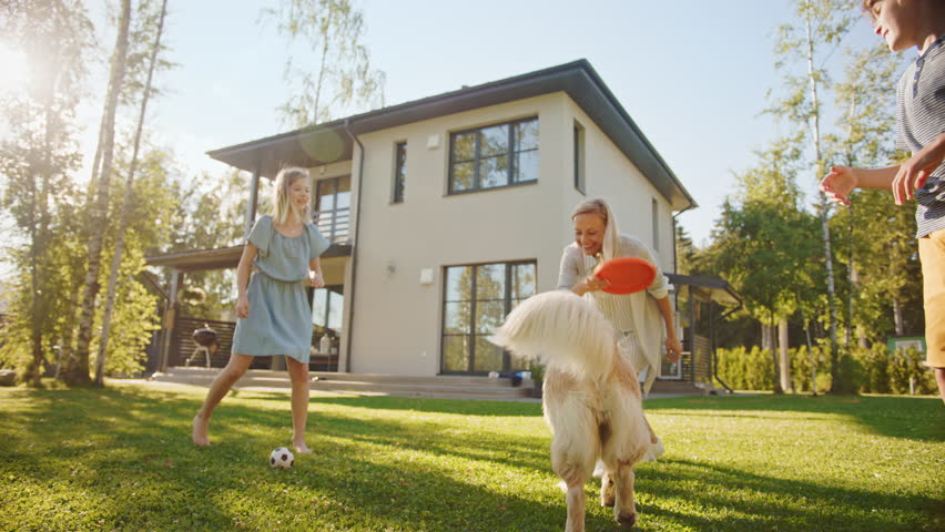 Smiling Beautiful Family of Four Play Catch flying disc with Happy Golden Retriever Dog on the Backyard Lawn. Idyllic Family Has Fun with Loyal Pedigree Dog Outdoors in Summer House. Handheld Low Shot