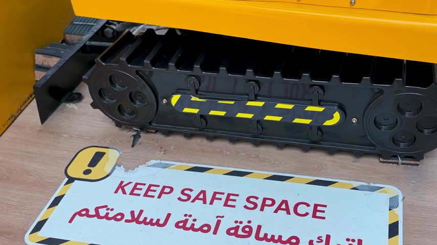 black caterpillar tracks on a yellow miniature toy excavator resting on an indoor wooden floor. A warning sticker reading Keep Safe Space in English and Arabic lies next to it.