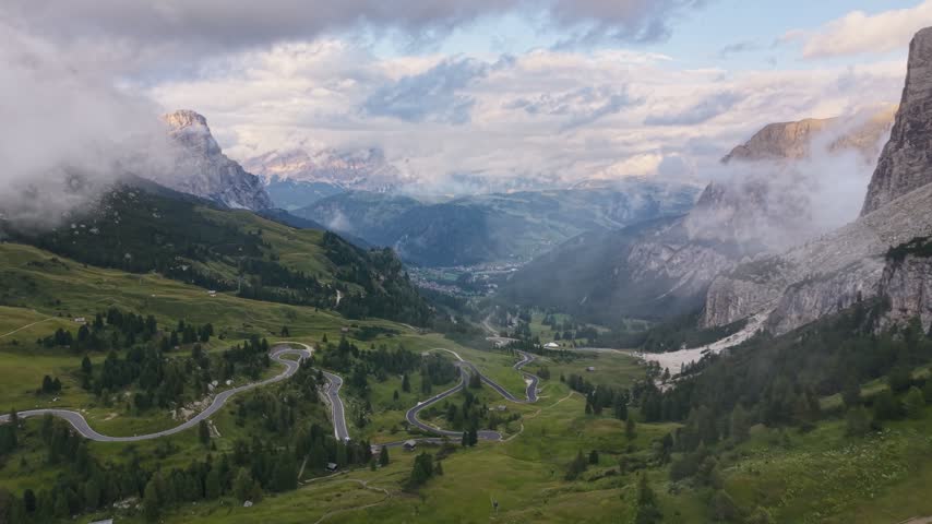 Cinematic drone flight over the winding mountain road at Gardena Pass in the Dolomites, revealing dramatic alpine valleys and rugged peaks as the camera passes through thin clouds and mist