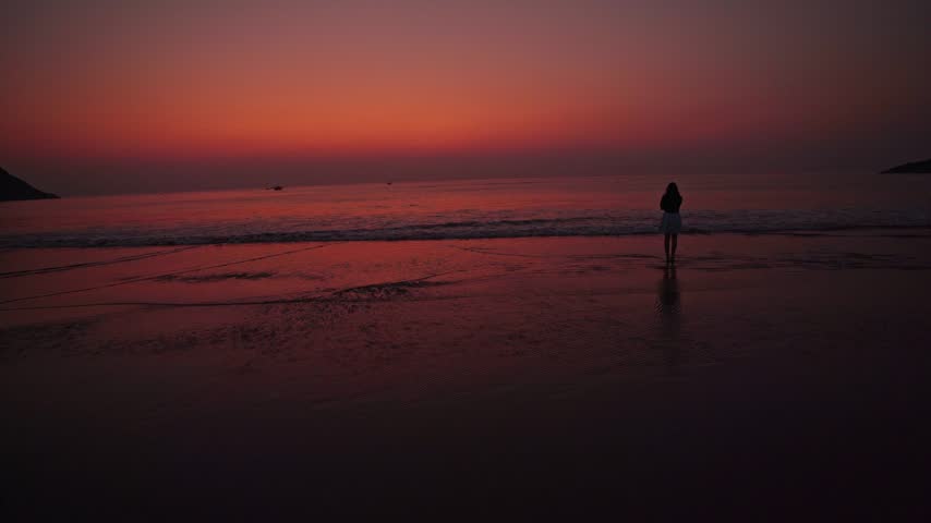 Silhouette of a woman standing at the seashore as gentle waves touch her feet during a peaceful evening sunset, evoking solitude, freedom, reflection, and emotional calm.