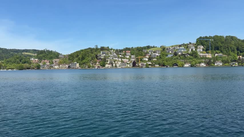 Scenic footage of Lucerne, Switzerland, captures riverside houses with the Alps extending across the background, creating a recognizable Swiss urban landscape. The composition brings together residential buildings, waterfront scenery, town architecture, and mountain surroundings, making it suitable for tourism campaigns, travel documentaries, destination guides, European vacation content, Swiss city features, and landscape productions.