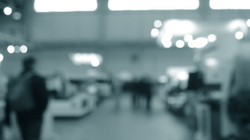 Blurred background. Many people walking inside large building. Men and women walking on floor inside room with high ceiling and large lanterns on walls. Public place. Exhibition presentation backdrop