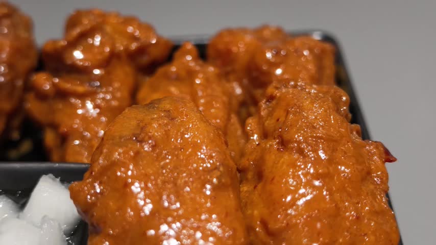 Extreme close up of crispy fried chicken wings coated in glossy spicy red sauce served with pickled radish in a black tray