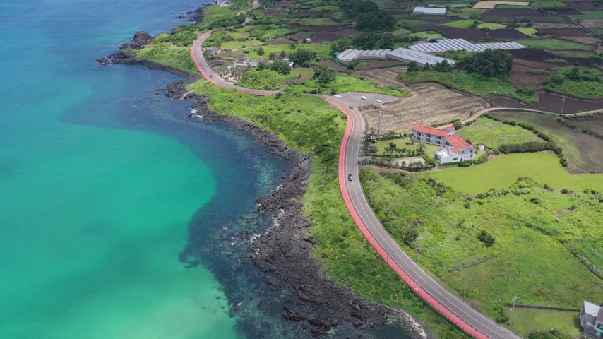 Horizontal cameo shot dollying forward with a downward tilt over Gujwa-eup, Jeju Island, South Korea, showing a coastal road, turquoise water, volcanic rocks, farmland and greenhouses