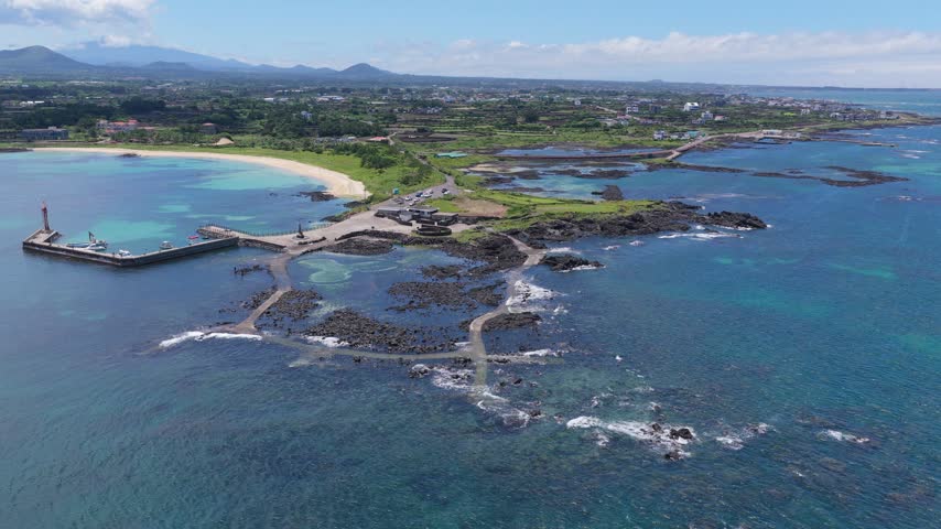 Horizontal cameo shot trucking right while facing left over Hado-ri, Jeju Island, South Korea, showing turquoise water, rocky coast, beach, harbor, farmland and mountains