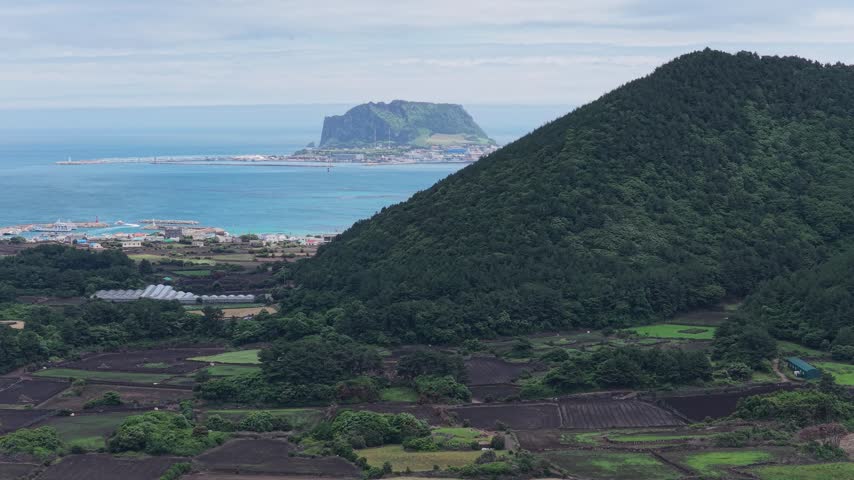 Horizontal cameo shot trucking left along Gujwa-eup, Jeju Island, South Korea, showing farmland, coastal village, volcanic shoreline, green hills and a distant volcanic island