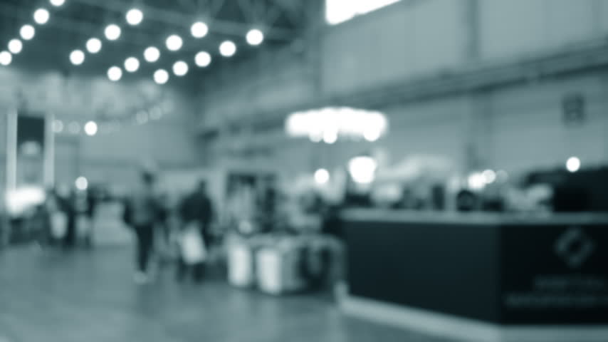 Blurred background. Many people walking inside large building. Men and women walking on floor inside room with high ceiling and large lanterns on walls. Public place. Exhibition presentation backdrop