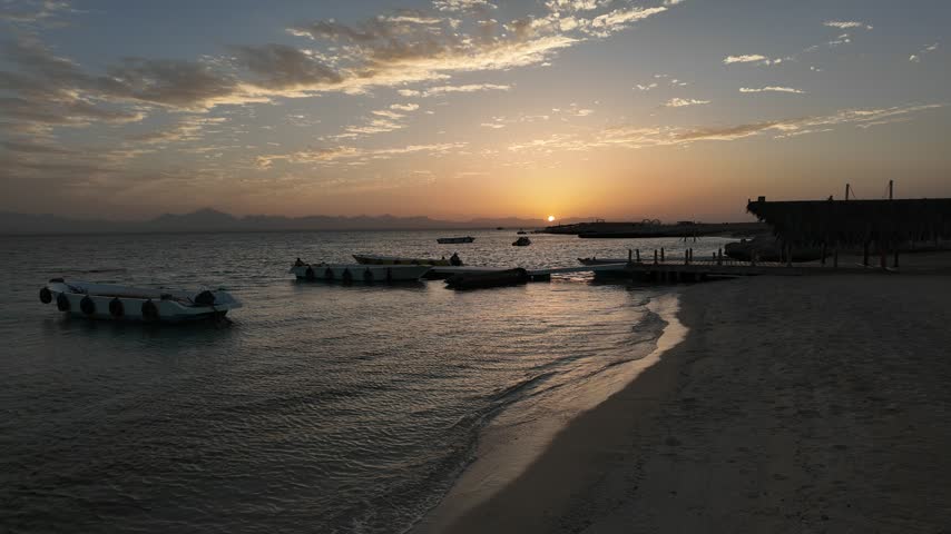 Beautiful sunset over the Red Sea at Eden Island near Hurghada, Egypt. Small boats float close to the sandy island shoreline as gentle waves wash onto the beach and warm golden sunlight reflects across the water beneath dramatic evening clouds. Scenic tropical seascape at a popular Red Sea island destination.