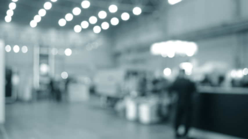 Blurred background. Many people walking inside large building. Men and women walking on floor inside room with high ceiling and large lanterns on walls. Public place. Exhibition presentation backdrop