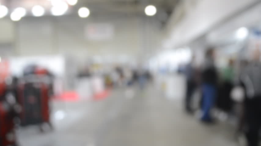 Blurred background. Many people walking inside large building. Men and women walking on floor inside room with high ceiling and large lanterns on walls. Public place. Exhibition presentation backdrop