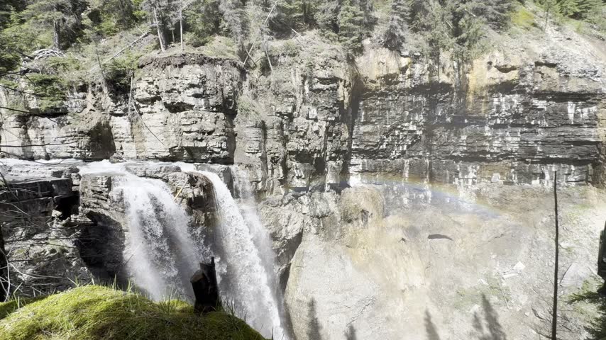 Johnston Creek river in Johnston Canyon in the Banff National Park. British Columbia, Canada. Waterfall flows down rocky cliffside, creating mist and a rainbow in the air, with trees framing the scene