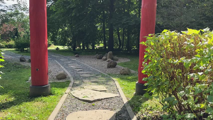 Traditional red torii gates border stone path in Japanese garden. Cobblestone walkway leads toward oriental pagoda house surrounded by trees. Perspective view frames Asian landscape architecture.