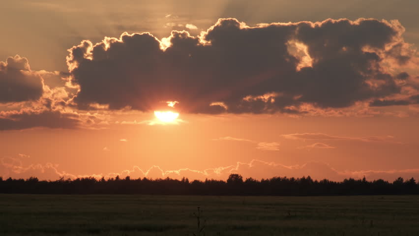 Picturesque cloudy landscape at sunset. Half sun shines under cloud in sky above field. Sunlight illuminates beautiful clouds over forest