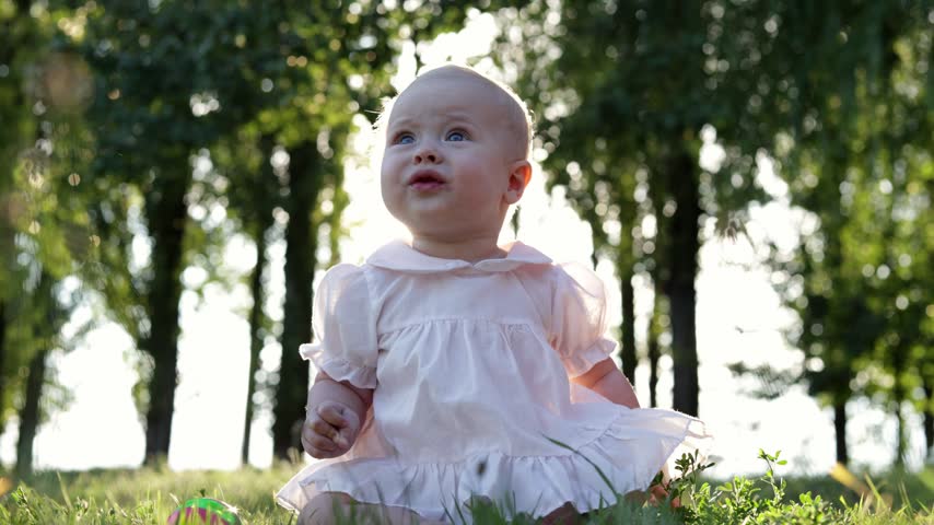 A curious baby girl with bright blue eyes looks up in wonder while sitting in a sunlit grassy field with trees in the background