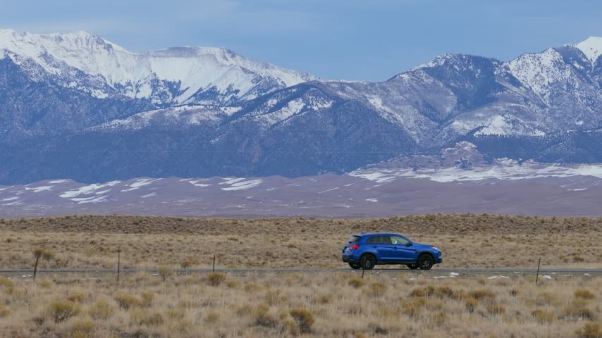 Aerial view of a blue car driving on a road through the desert landscape with snow-capped mountains in the background near the Great Sand Dunes, Alamosa, Colorado, United States.