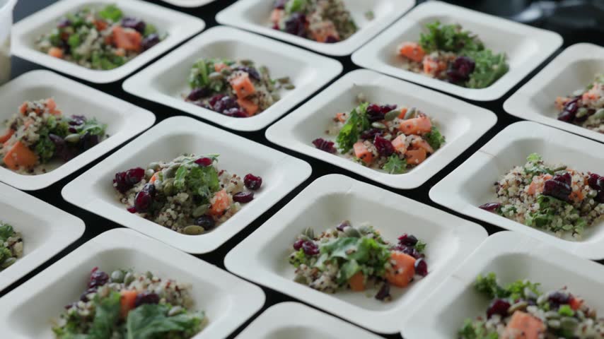 Rows of tri-color quinoa salad with kale, roasted sweet potato, dried cranberries and pumpkin seeds portioned into white square plates on a black table, photographed at a low angle