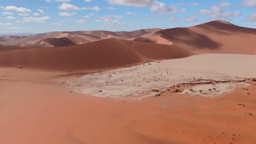 Aerial view of Deadvlei in Sossusvlei, Namib Desert, Namibia, with dead camelthorn trees, orange dunes, tiny people, and faint tracks in clear daylight with soft shadows.