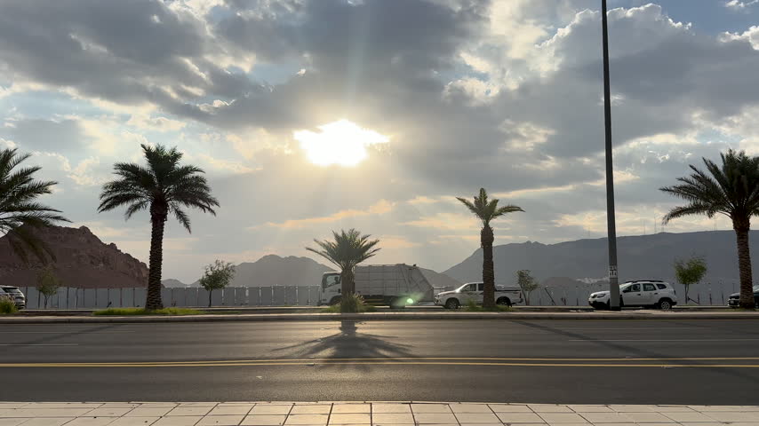Cars drive along a paved road lined with palm trees against a mountain background under sun rays through clouds.