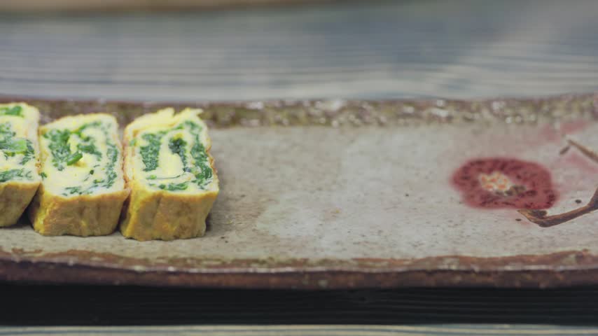 A close-up shot of hands arranging freshly sliced Korean rolled omelette (Gyeran-mari) onto a vintage ceramic plate. 