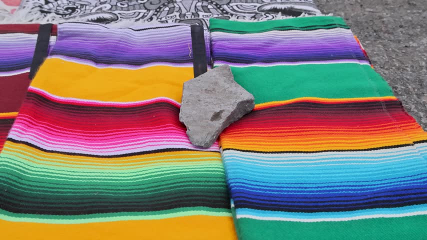 Brightly striped Mexican serape blankets displayed outdoors with a stone weight at a Mexico City market.