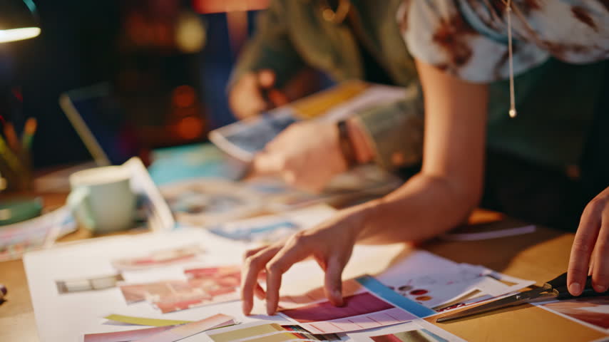 Hands closeup cutting magazine photos with scissors, arranging mood board on wooden desk. Two coworkers collaborating in cozy evening studio, focused creative teamwork in warm lamp light lifestyle.