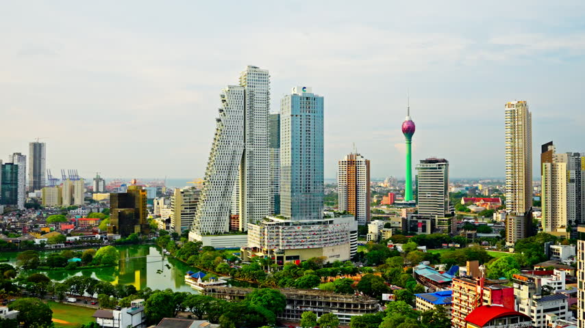 Wide panorama of the Colombo skyline, Sri Lanka, sweeping from the twin Altair towers and downtown skyscrapers to the Lotus Tower, with green parks and lake waters woven through the city.