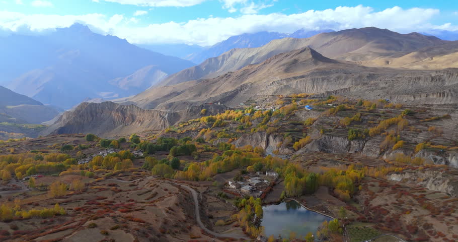 Annapurna Circuit, Mustang Nepal, Aerial shot of a small reflective pond, stone houses, golden autumn trees, and terraced fields set against rugged desert hills and towering Himalayan peaks.
