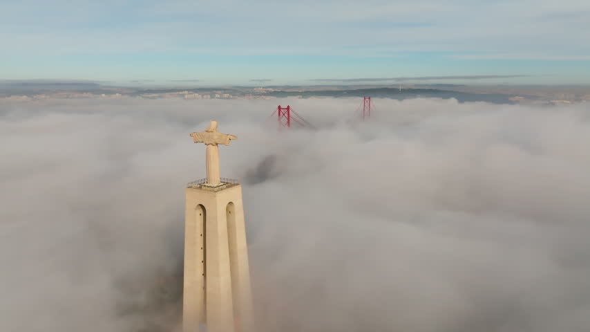 Drone flies toward Cristo Rei statue above dense fog, Lisbon, Almada. 25 de Abril Bridge top emerges from mist at sunrise. Cinematic Portugal aerial footage