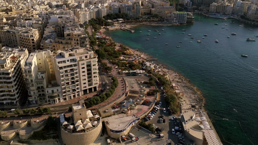 Drone flies along Malta bayfront with apartments, rocky shoreline, parked cars and boats in calm water.