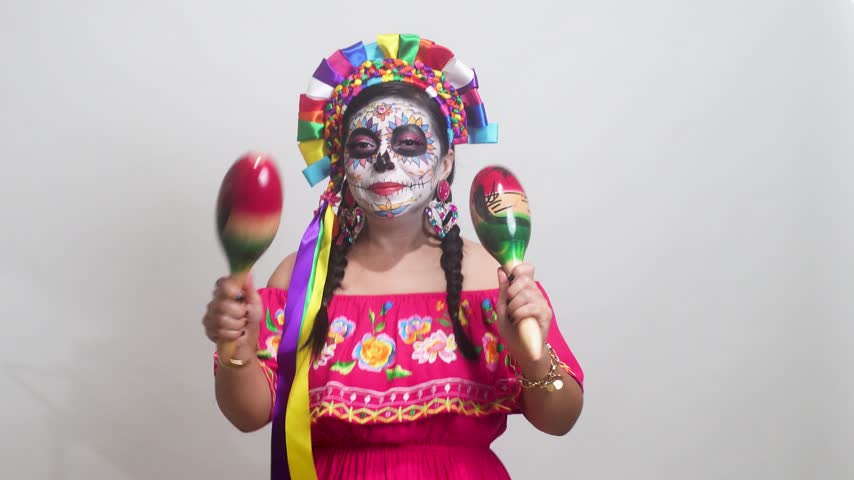 Mexican woman with Catrina makeup and traditional dress happily plays maracas against a clean white background.