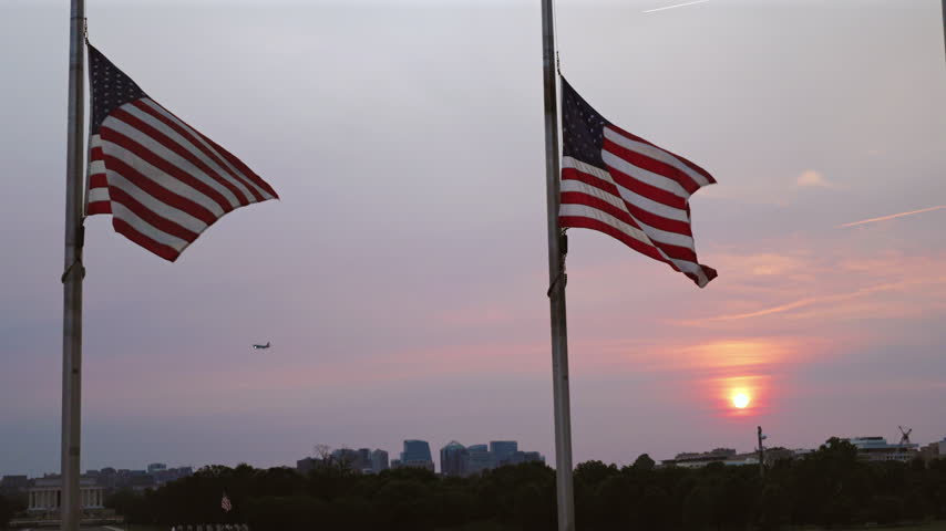 Two American flags fly at half-staff on flagpoles as sun sets over Washington DC skyline. Pink and orange hues fill sky while an airplane approaches in distance and city buildings create a silhouette.